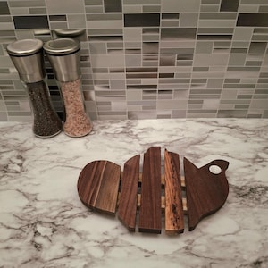 May include: A wooden trivet in the shape of a fish, with a mix of dark and light brown wood slats. The trivet is on a marble countertop, with two stainless steel salt and pepper grinders in the background.