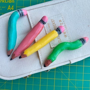 Könnte beinhalten: Vier farbige Bleistift-Figuren, zwei sind gebogen und zwei sind gerade. Die Figuren sind türkis, rosa, gelb und grün. Die Figuren befinden sich auf einem weißen Canvas-Hintergrund mit einer grünen Schneidematte.