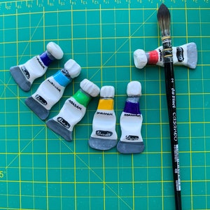 Puede incluir: Una colección de imanes de cerámica con forma de tubos de pintura en varios colores, incluyendo azul, verde y amarillo, dispuestos sobre una esterilla de corte verde. Un pincel negro da Vinci Casaneo descansa entre los imanes.