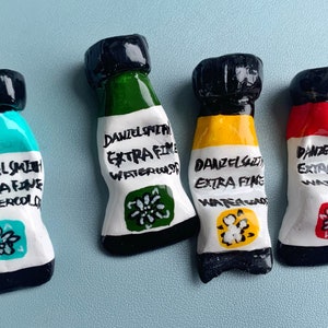 Puede incluir: Cuatro tubos de pintura de acuarela en miniatura de colores. Cada tubo está etiquetado como "DANIEL SMITH EXTRA FINE WATERCOLOR" con un color único: turquesa, verde, amarillo y rojo. Los tubos tienen tapas negras y etiquetas blancas con diseños florales.