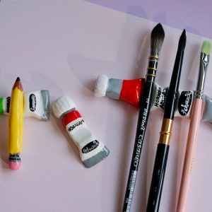 May include: Ceramic brush rests shaped like paint tubes, including one with a yellow pencil design. The white, red, and grey tubes feature black text labels. Three artist paintbrushes are resting across the ceramic holders, showcasing varied bristles and handles in black and light pink.