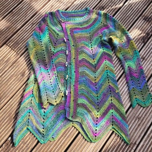 Op de afbeelding: Een veelkleurige gehaakte cardigan met een chevron-patroon en een knoopsluiting. De cardigan is gemaakt met verschillende tinten groen, blauw, paars en roze garen.