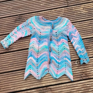 Może przedstawiać: Ręcznie robiony kardigan z dzianiny w chevronowy wzór w odcieniach niebieskiego, różowego i szarego. Kardigan zapinany na guziki, z lekko rozszerzanym dołem.