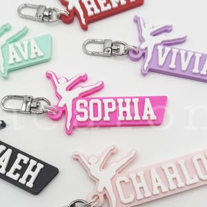Puede incluir: Una colección de llaveros personalizados con una silueta de bailarina y nombres como "Remy" y "Ava". Los llaveros vienen en varios colores, incluyendo rojo, verde menta y rosa, con cierres plateados.