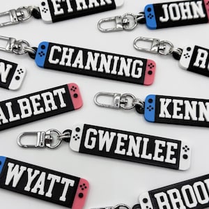 May include: A collection of personalized keychains with names like Ethan, John, and Jen. Each keychain features a black rectangular design with white lettering and colorful accents, including blue and pink details, and a silver clasp.