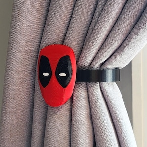 May include: A red and black 3D printed Deadpool mask curtain tieback. The tieback is attached to a grey curtain.