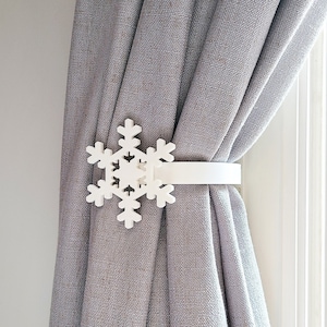May include: White snowflake curtain tieback with a white strap. The tieback is attached to a gray curtain.