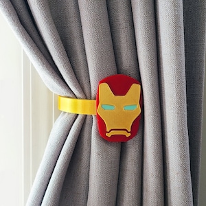 May include: A yellow and red curtain tieback featuring a superhero mask design. The tieback is attached to a grey curtain.