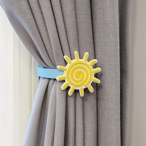 May include: A blue and yellow curtain tieback with a sun design. The tieback is attached to a gray curtain.