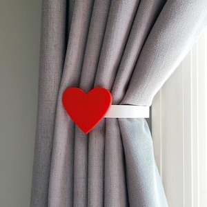 May include: A red heart-shaped curtain tieback with a white band, holding a gray curtain. The tieback is made of a solid material and is positioned near a window, adding a decorative touch to the window treatment.