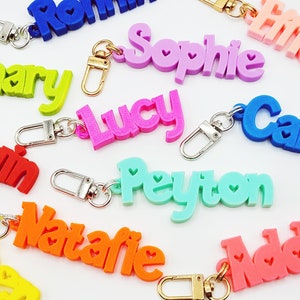 May include: A collection of colorful personalized keychains with names like Sophie, Lucy, Peyton, and Natalie. Each keychain features a unique name in bold, 3D lettering with small heart accents.