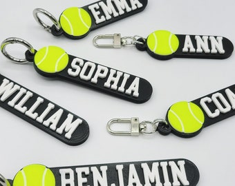 May include: Five black and white personalized keychains with a yellow tennis ball design. The keychains feature the names "Emma", "Ann", "Sophia", "William", and "Benjamin".
