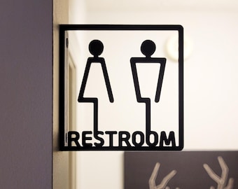 May include: Black metal sign with a silhouette of a woman and a man, indicating a restroom. The sign reads "RESTROOM".
