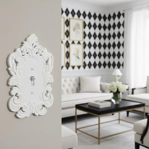 May include: A white, ornate light switch cover with a decorative, scroll-like design. The cover is mounted on a light gray wall. The switch is in the on position. The background shows a living room with a black and white diamond pattern wall.