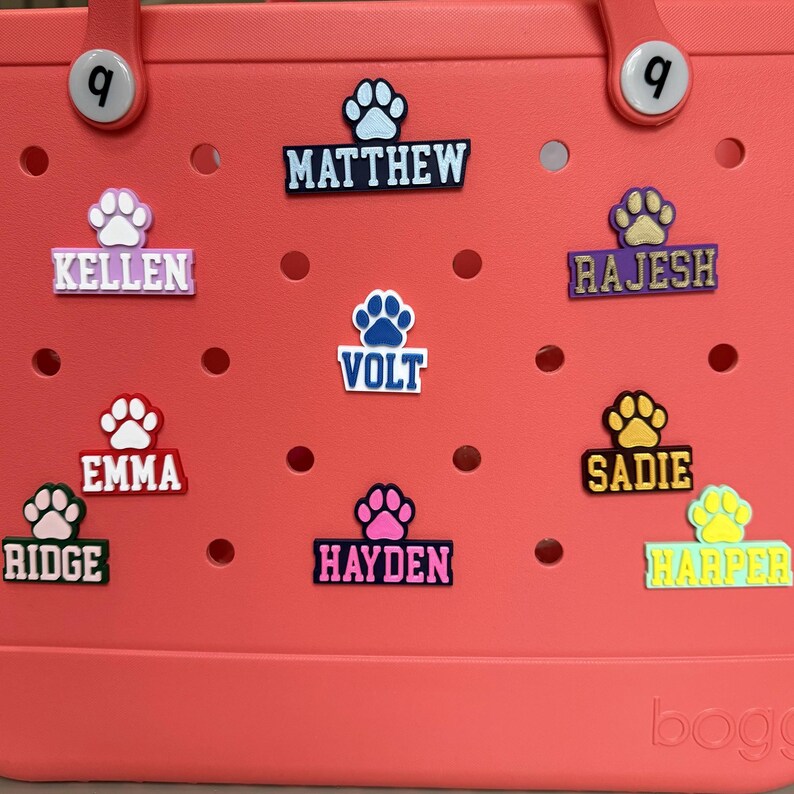 May include: A pink and white tote bag with various colorful paw print charms. The charms have names on them, including "Kellen", "Matthew", "Volt", "Emma", "Ridge", "Hayden", "Rajesh", "Sadie", and "Harper".