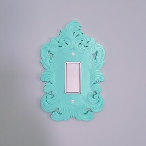 May include: A decorative light switch cover in a vibrant turquoise color. The cover features an ornate, swirling design around a rectangular opening for the switch. The switch itself is white, and the cover is mounted on a gray wall.