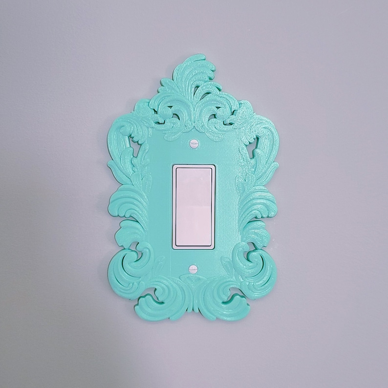 May include: A decorative light switch cover in a vibrant turquoise color. The cover features an ornate, swirling design around a rectangular opening for the switch. The switch itself is white, and the cover is mounted on a gray wall.