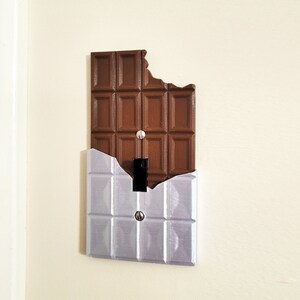 Puede incluir: Una tapa de interruptor de luz con forma de barra de chocolate con un mordisco. La tapa es de pl&aacute;stico marr&oacute;n y tiene una placa de metal plateado detr&aacute;s.
