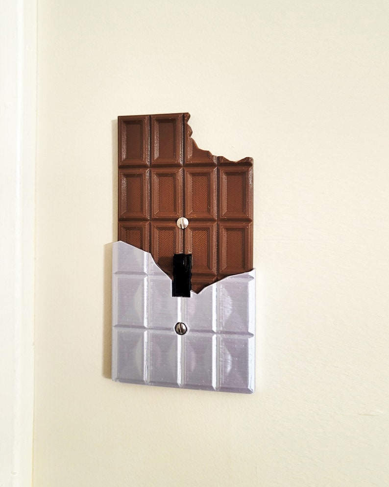 Puede incluir: Una tapa de interruptor de luz con forma de barra de chocolate con un mordisco. La tapa es de pl&aacute;stico marr&oacute;n y tiene una placa de metal plateado detr&aacute;s.