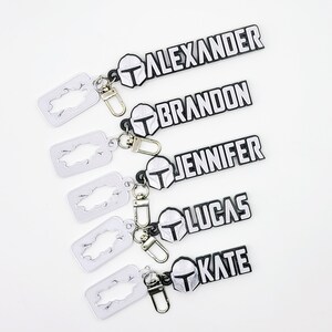 May include: Five black and white keychains with silver split rings. Each keychain features a Mandalorian helmet and a name: Alexander, Brandon, Jennifer, Lucas, and Kate.