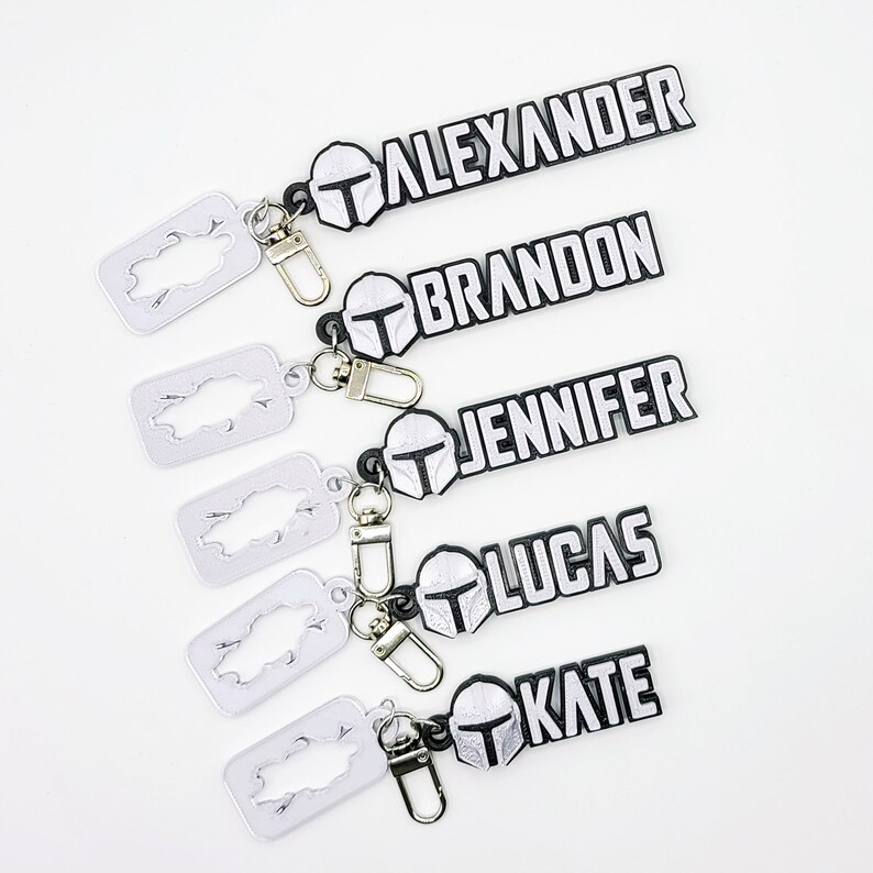 May include: Five black and white keychains with silver split rings. Each keychain features a Mandalorian helmet and a name: Alexander, Brandon, Jennifer, Lucas, and Kate.
