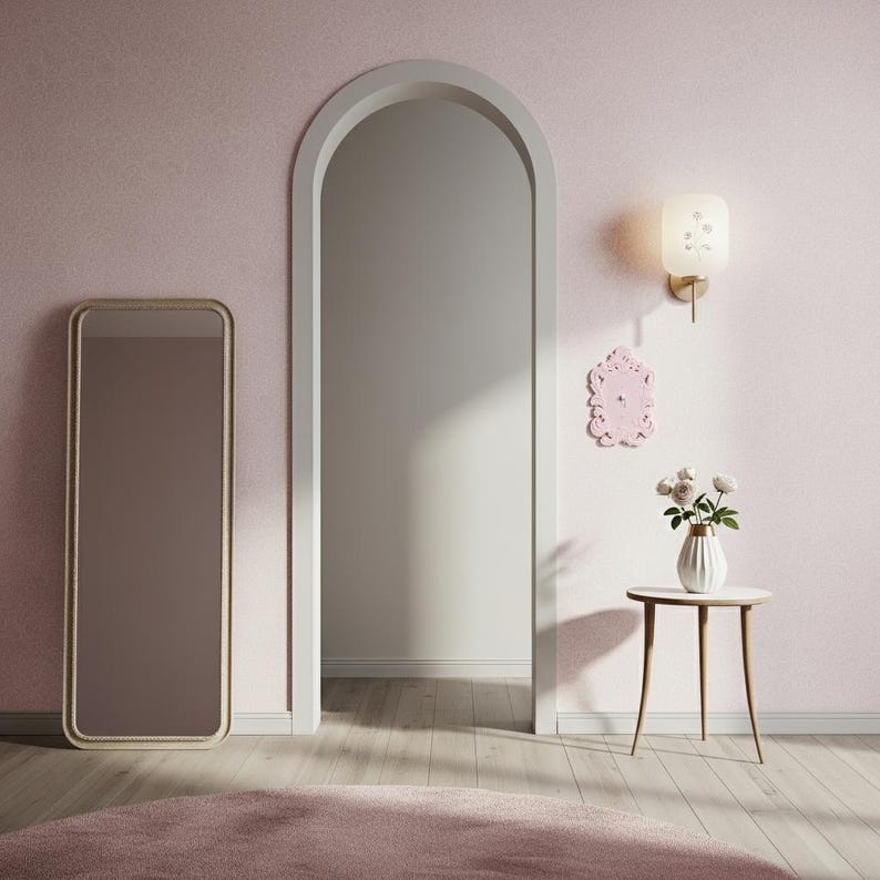 May include: A room with a pink wall, a large arched doorway, and a full-length mirror with a gold frame. A small side table holds a vase of white flowers. A pink rug and a wall sconce complete the decor.