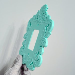 May include: A decorative, light teal switch plate with an ornate, swirling floral design. The rectangular plate has a single rectangular opening for a light switch and two screw holes. The plate is held by a gloved hand.