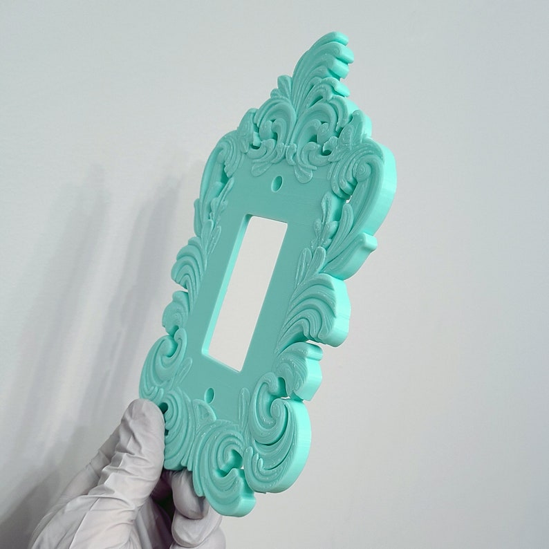 May include: A decorative, light teal switch plate with an ornate, swirling floral design. The rectangular plate has a single rectangular opening for a light switch and two screw holes. The plate is held by a gloved hand.