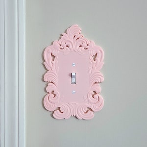 May include: A light pink, ornate light switch cover with a decorative frame. The cover has a single toggle switch and is mounted on a light gray wall. The frame features a detailed, swirling design.