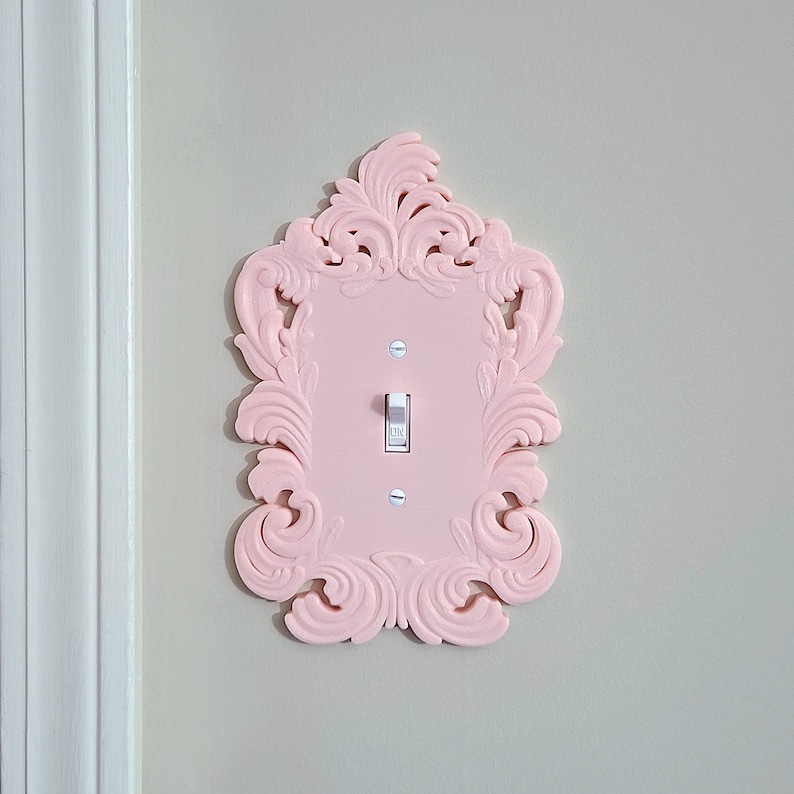 May include: A light pink, ornate light switch cover with a decorative frame. The cover has a single toggle switch and is mounted on a light gray wall. The frame features a detailed, swirling design.