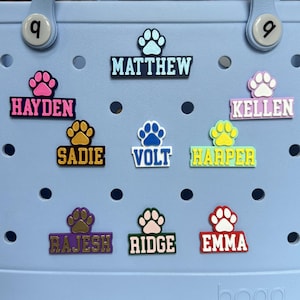 May include: Nine colorful personalized dog paw print name tags with a paw print design. The names are: Hayden, Matthew, Kellen, Sadie, Volt, Harper, Rajesh, Ridge, and Emma.