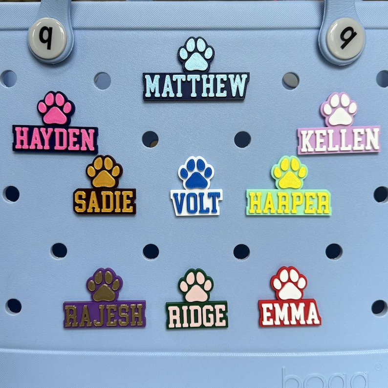 May include: Nine colorful personalized dog paw print name tags with a paw print design. The names are: Hayden, Matthew, Kellen, Sadie, Volt, Harper, Rajesh, Ridge, and Emma.