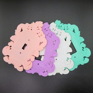 May include: A collection of decorative light switch covers in pastel colors: pink, purple, white, and mint green. Each cover features an ornate, swirling design. The covers are made of a solid material and are arranged on a dark surface.