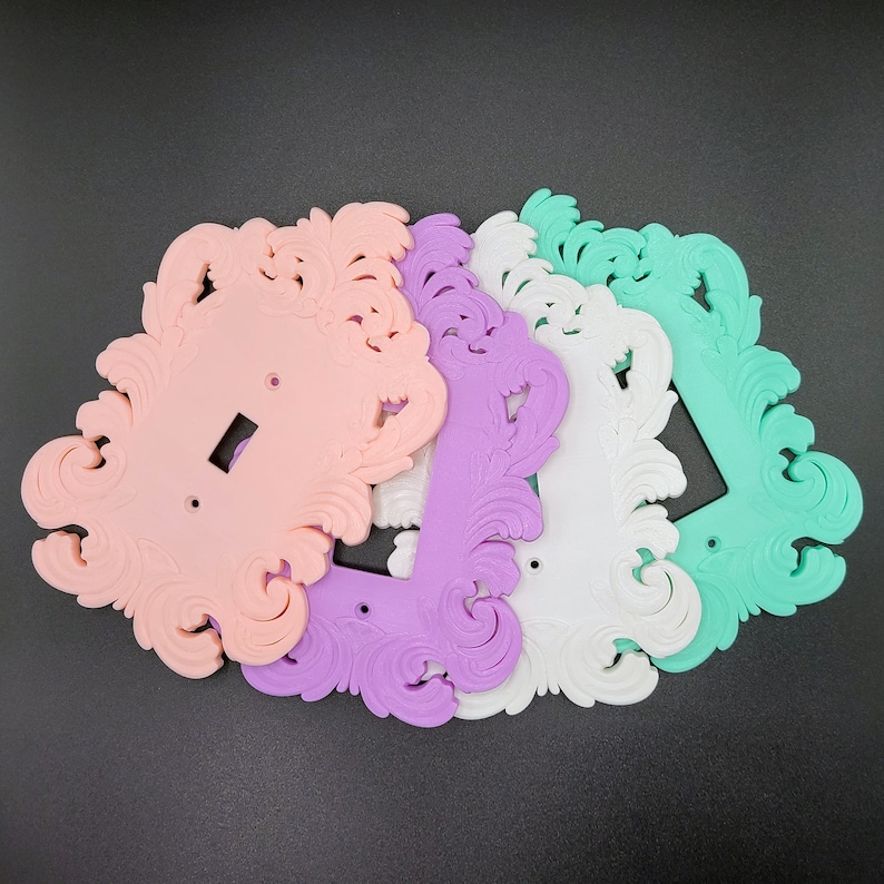 May include: A collection of decorative light switch covers in pastel colors: pink, purple, white, and mint green. Each cover features an ornate, swirling design. The covers are made of a solid material and are arranged on a dark surface.