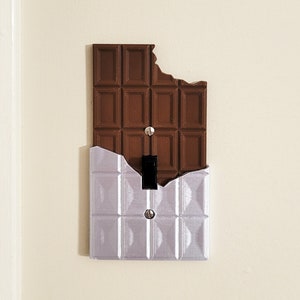 Puede incluir: Una tapa de interruptor de luz con forma de barra de chocolate con un mordisco. La mitad superior de la tapa es marr&oacute;n y la mitad inferior es plateada.