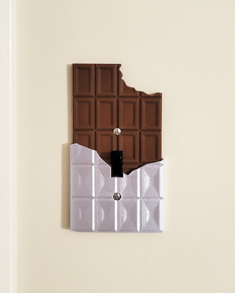 Puede incluir: Una tapa de interruptor de luz con forma de barra de chocolate con un mordisco. La mitad superior de la tapa es marr&oacute;n y la mitad inferior es plateada.