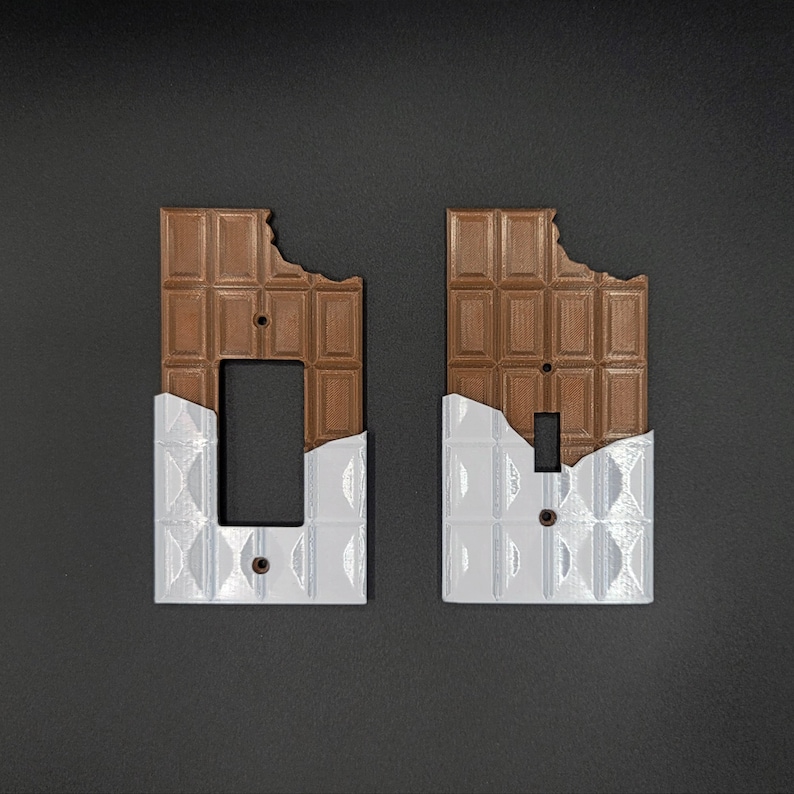 Puede incluir: Dos tapas de interruptor de luz con un dise&ntilde;o de barra de chocolate. El chocolate es marr&oacute;n y tiene un mordisco. Las tapas de los interruptores son blancas con un dise&ntilde;o texturizado.