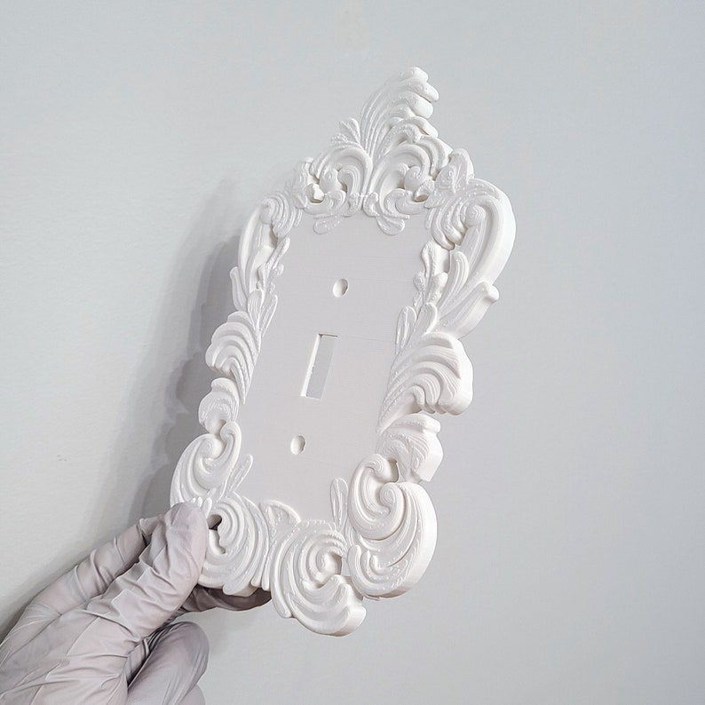 May include: A white, ornate light switch cover with a decorative, swirling floral design. The cover has a single switch opening and two screw holes. The design is a classic, elegant style.