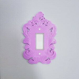 May include: A decorative light switch cover in a vibrant lavender hue. The cover features an ornate, swirling design around a rectangular opening for the switch. The switch itself is white, contrasting with the lavender frame. The cover is mounted on a light gray wall.