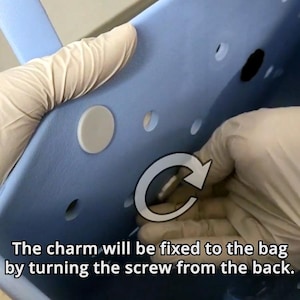 May include: A close-up of a light blue tote bag with a white screw and a white circle. The screw is being turned clockwise to attach a charm to the bag.