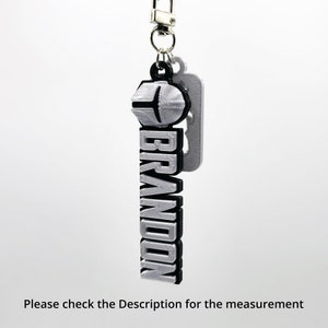 May include: A silver keychain with a black outline and the word "BRANDON" in bold letters. The keychain has a silver ring and a silver hook.