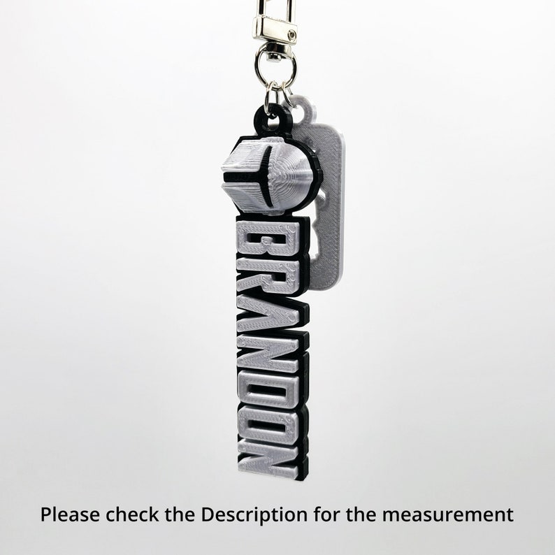 May include: A silver keychain with a black outline and the word "BRANDON" in bold letters. The keychain has a silver ring and a silver hook.