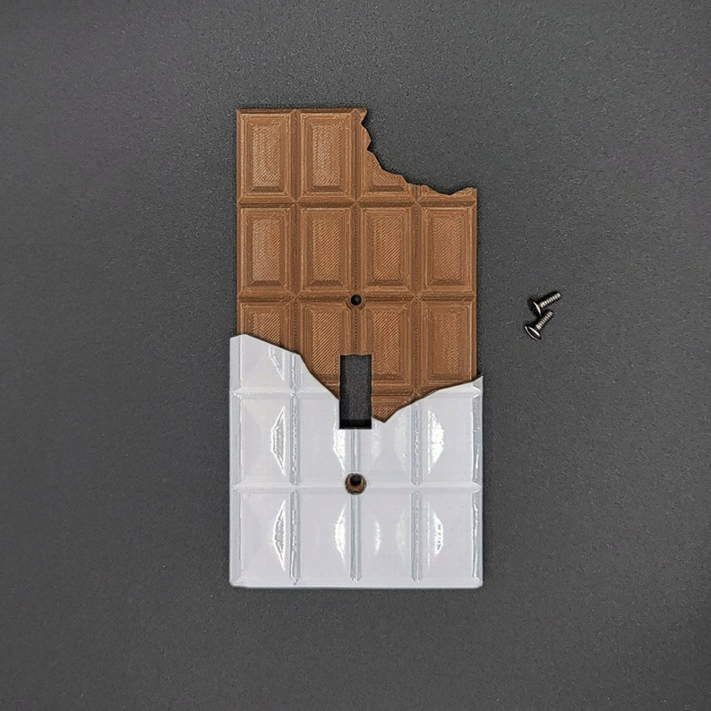 Puede incluir: Una tapa de interruptor de luz blanca con un dise&ntilde;o de barra de chocolate marr&oacute;n. La barra de chocolate tiene un mordisco.
