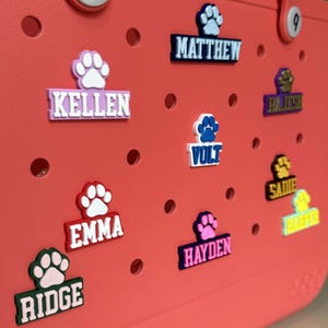 May include: A pink, red, green, blue, and purple set of personalized name tags with paw prints. The names on the tags are Kellen, Emma, Ridge, Volt, Hayden, Matthew, Sadie, and Harper.