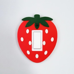 May include: A red strawberry-shaped light switch cover with white spots and green leaves. The cover has a white switch in the centre. The decorative cover is designed to fit over a standard light switch.