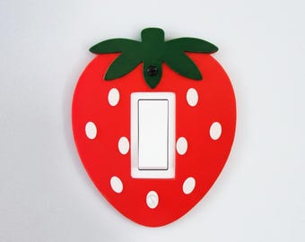 Pode incluir: Uma capa de interruptor de luz vermelha em forma de morango com bolinhas brancas e folhas verdes. A capa tem um interruptor branco no centro. A capa decorativa foi projetada para caber em um interruptor de luz padrão.