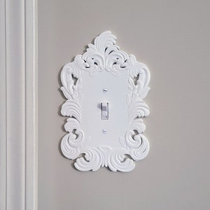 May include: White light switch cover with an ornate, scrollwork design. The cover has a rectangular center with a toggle switch and is mounted on a light gray wall. The decorative frame adds a touch of elegance.