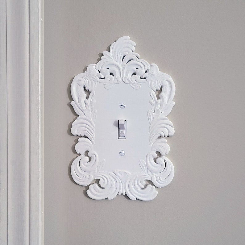 May include: White light switch cover with an ornate, scrollwork design. The cover has a rectangular center with a toggle switch and is mounted on a light gray wall. The decorative frame adds a touch of elegance.