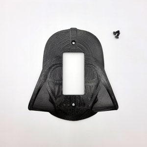 May include: Black, 3D-printed light switch cover featuring the likeness of Darth Vader's helmet. The cover has a rectangular opening for the switch and includes three small black screws for installation. The design is detailed and textured.