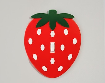 Pode incluir: Uma capa de interruptor em forma de morango vermelho com detalhes de sementes brancas e uma parte superior de folha verde. A capa tem um interruptor de luz branco no centro. A capa é montada numa parede cinza claro.